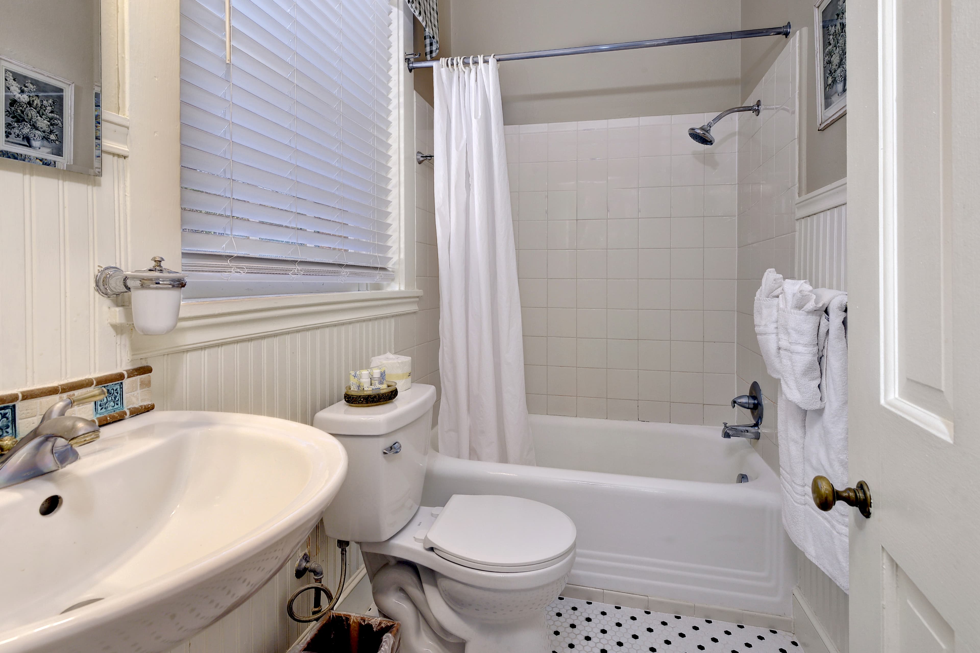 Bathroom with white wainscoting, textiles in shades of white, cream and blue, and white pedestal sink with decorative blue and tan backsplash tiles