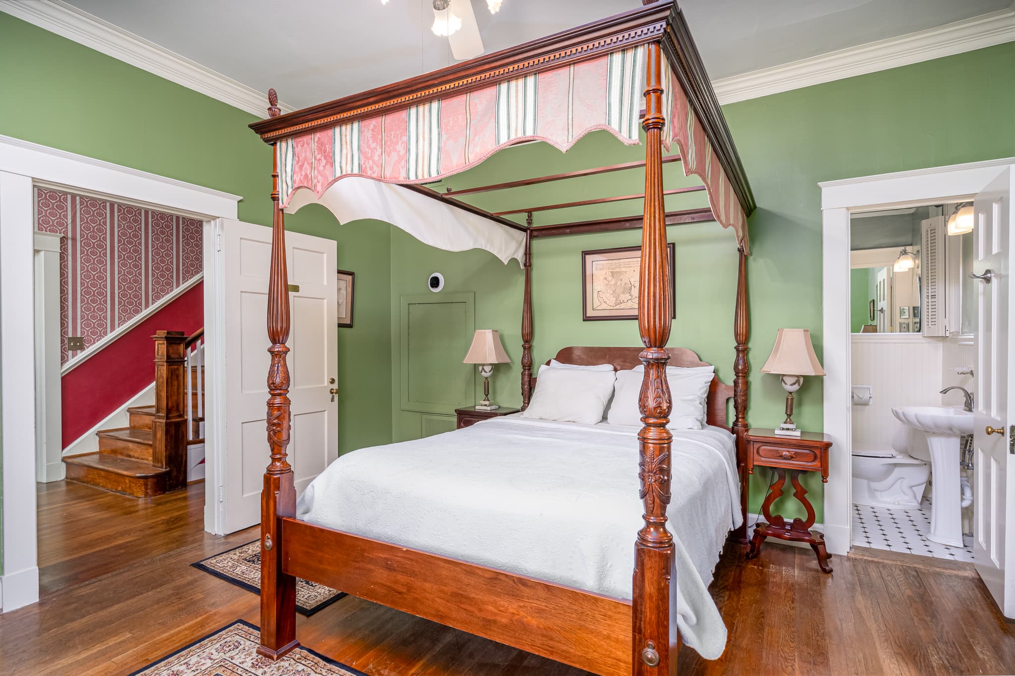 Carved mahogany four-poster bed with nightstands and lamps, hardwood floors, doorway to bathroom, and doorway leading to hallway with stairs