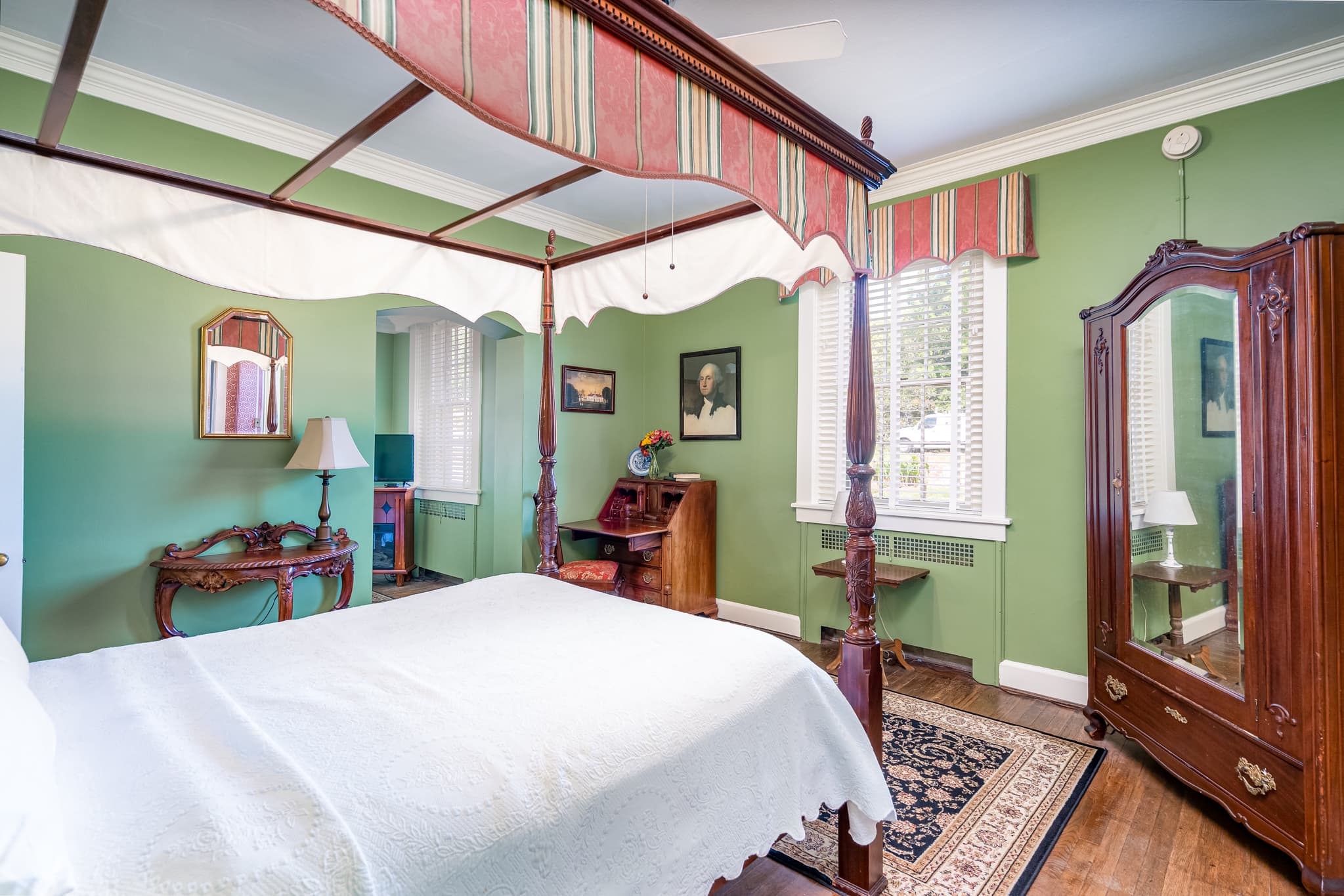 Bedroom with carved mahogany four-poster bed with nightstands and lamps, ornate oriental rug, antique armoire with full length mirror, and green painted walls