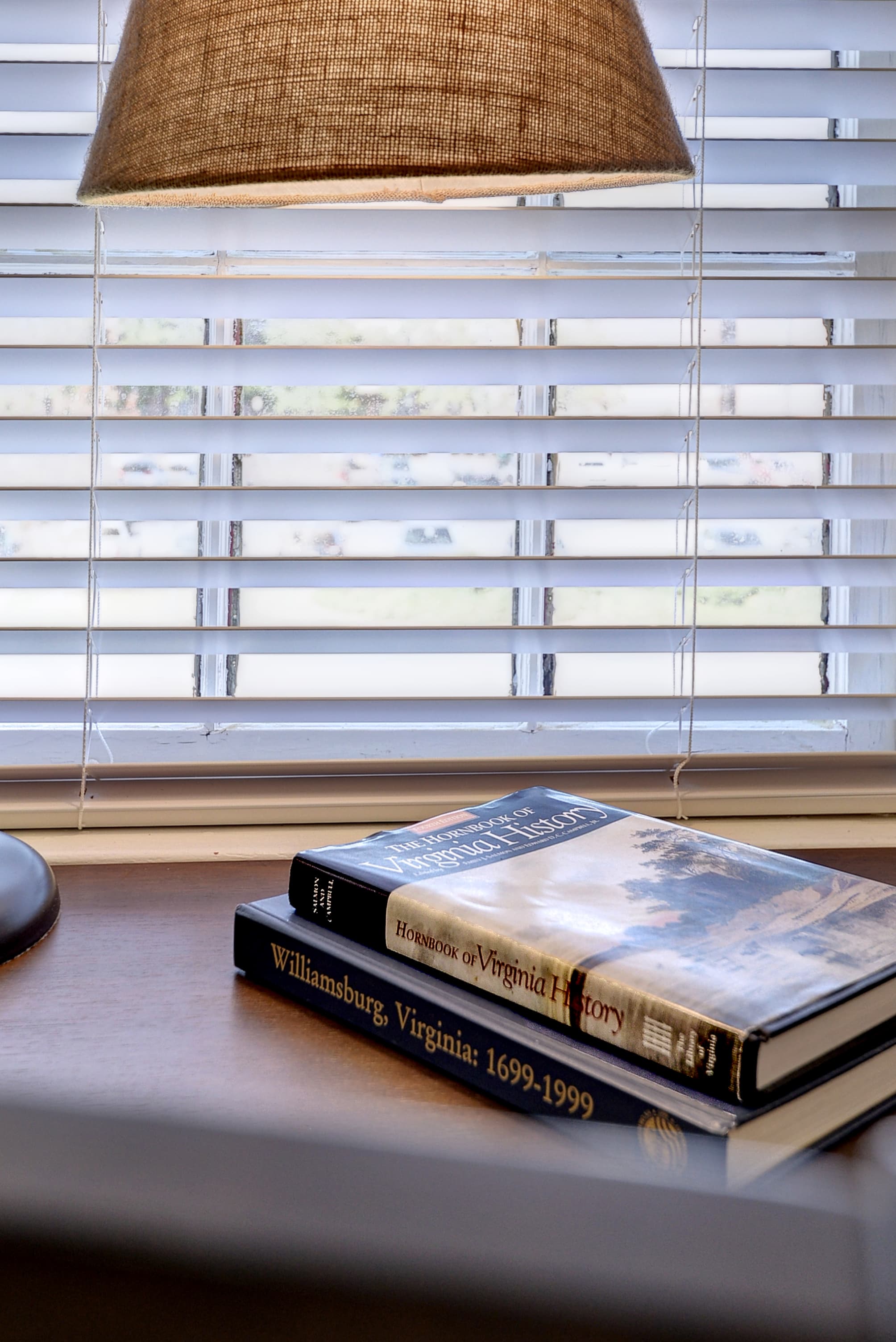 A lamp illuminates a stack of books on a desk beside a window with blinds.