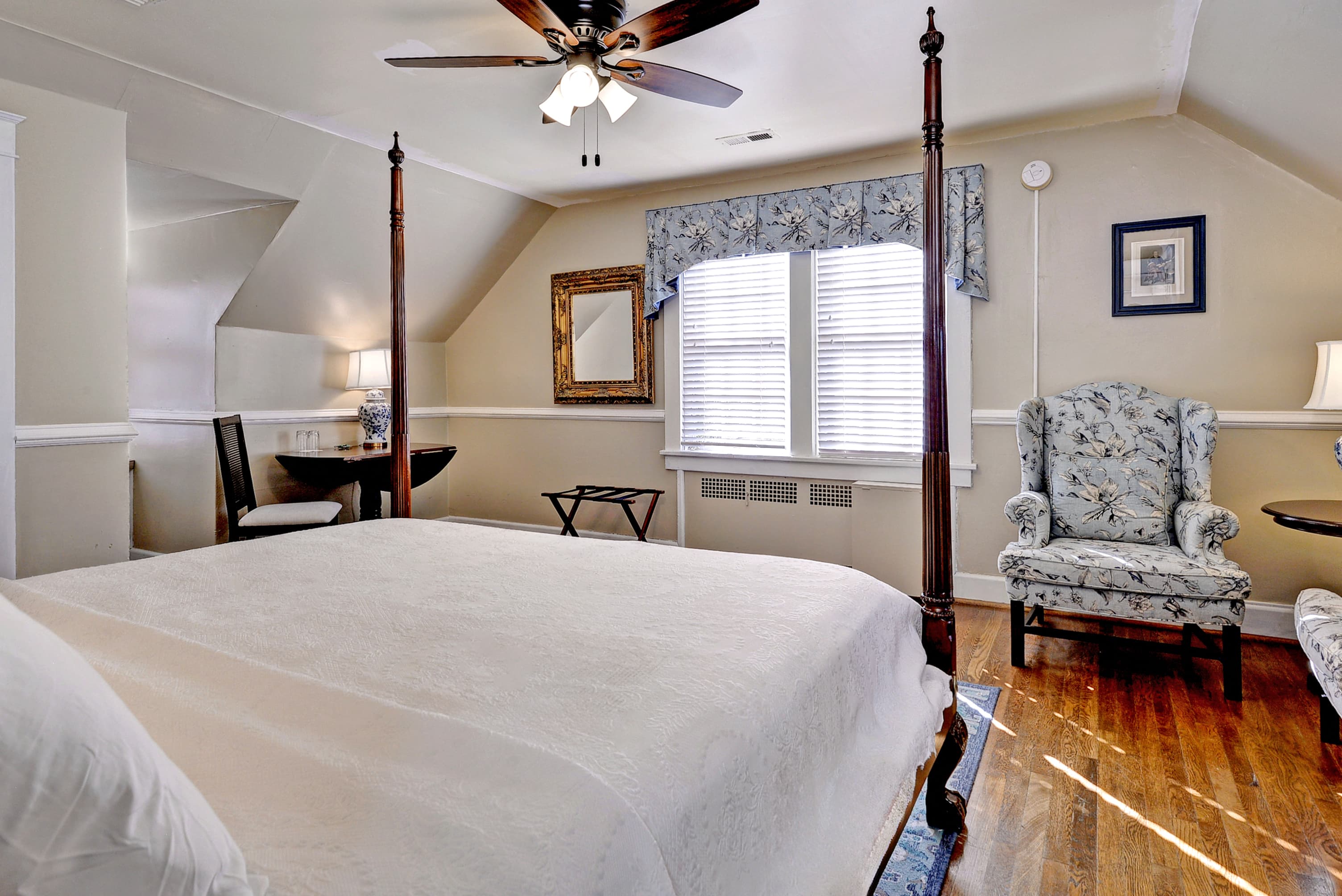 A cozy bedroom featuring a four-poster bed, a floral armchair, and a desk by a window.