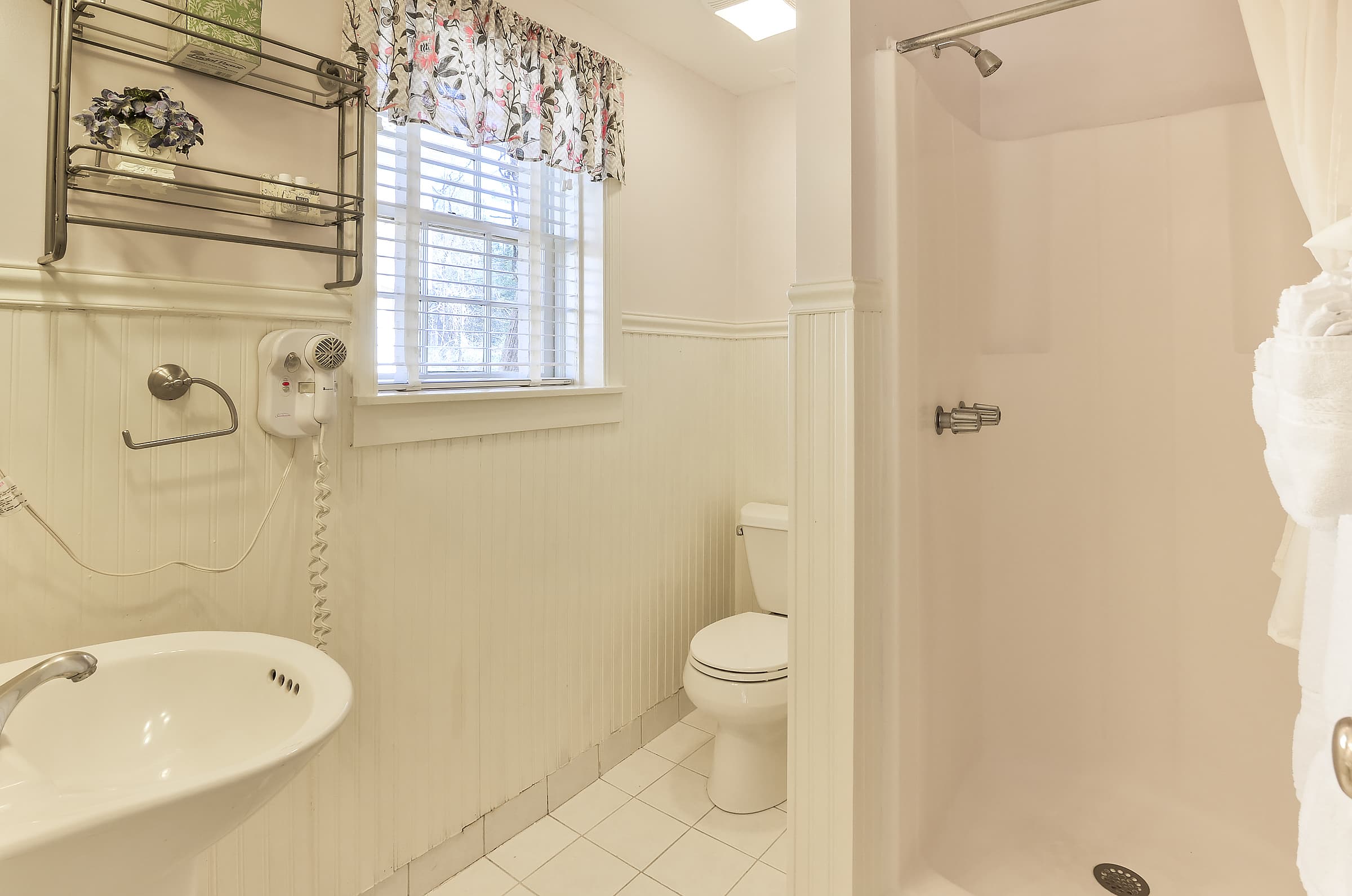 Small bathroom featuring a shower, toilet, sink, and a window with floral curtains.
