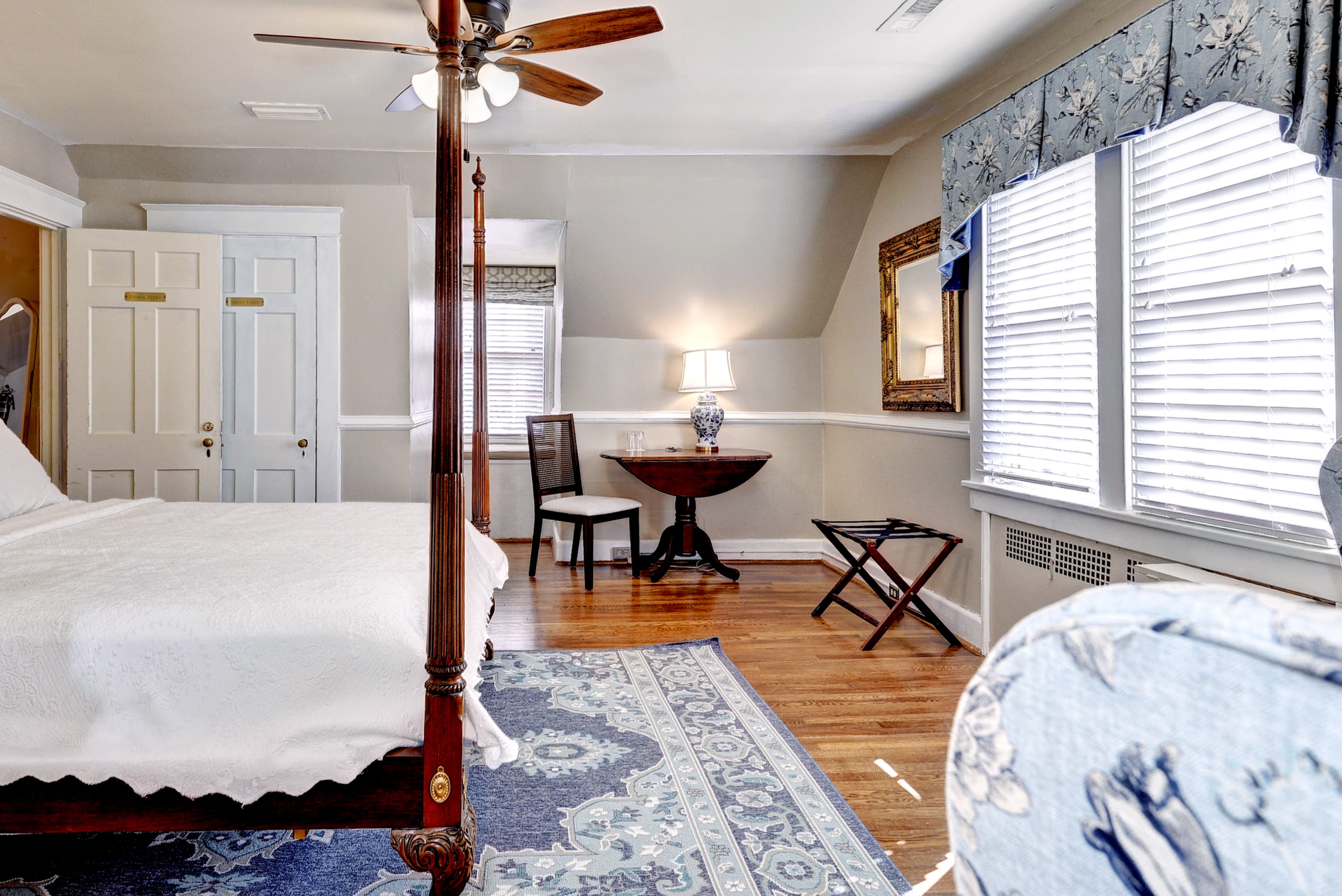 A cozy bedroom featuring a four-poster bed, a small round table with a lamp, and large windows with blue curtains.