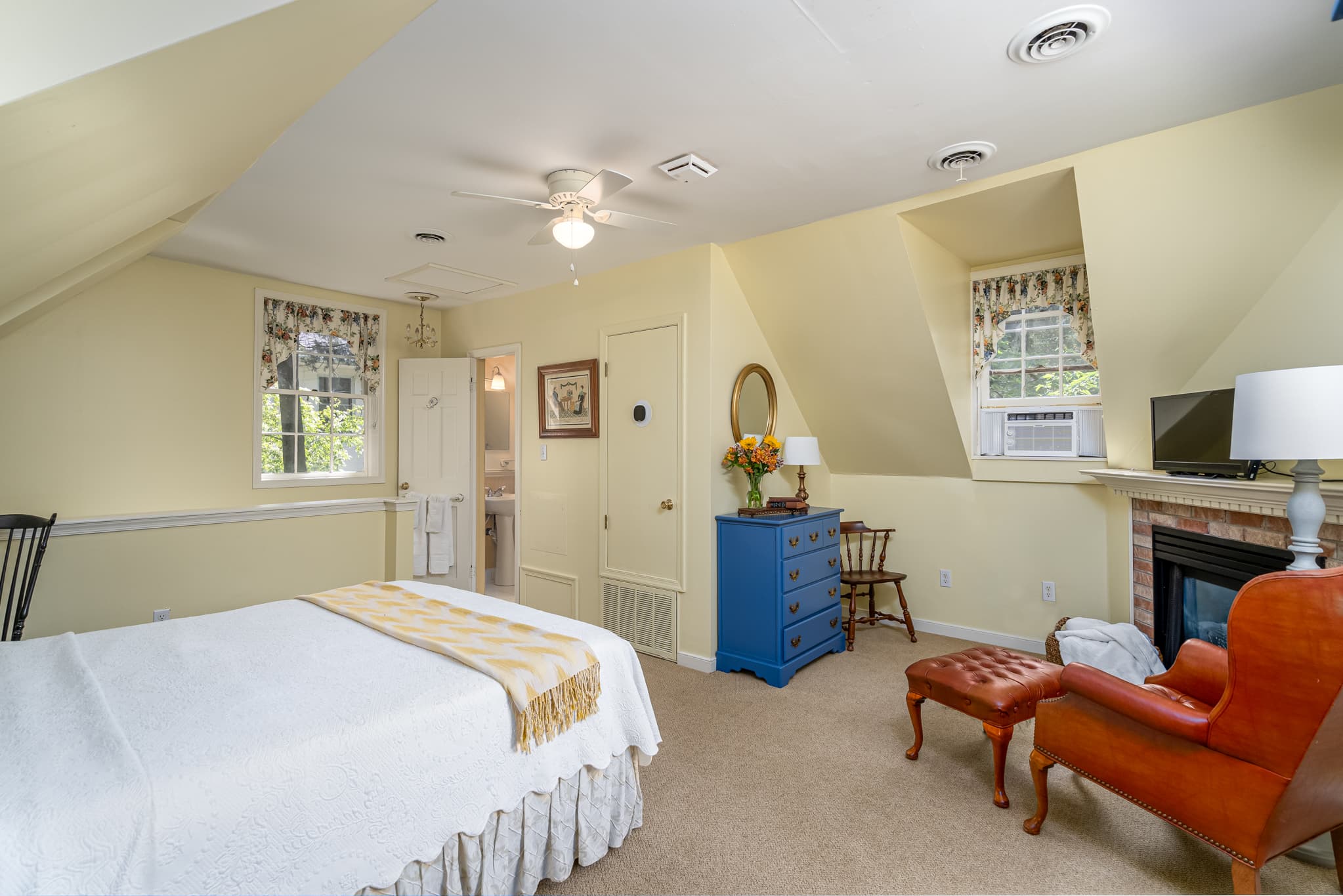 A cozy bedroom with a queen bed, vintage furniture, and natural light from windows in a softly colored room.