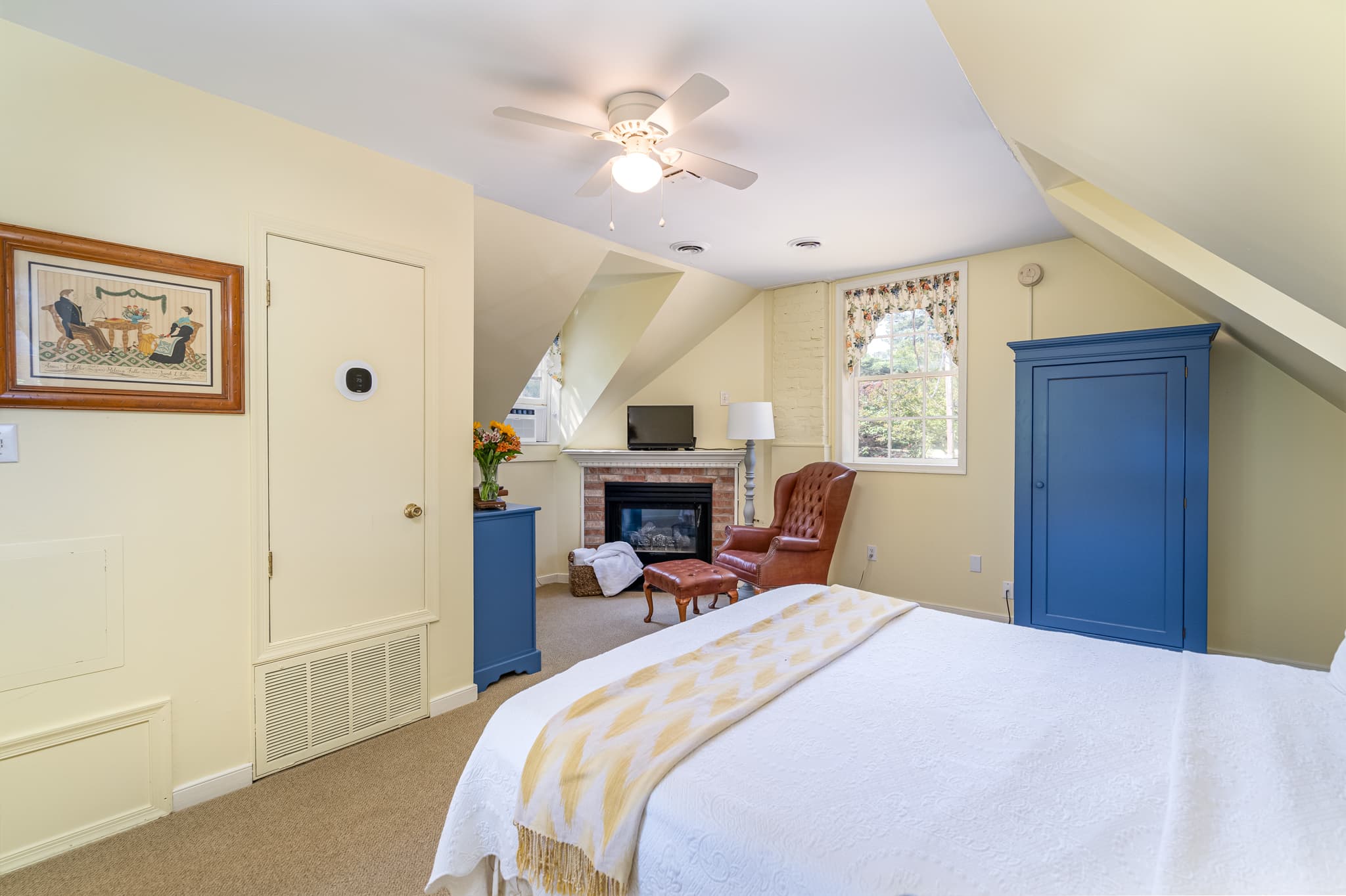 Bright, cozy bedroom with a queen bed, armchair, and fireplace, featuring sloped ceilings and decorative accents.