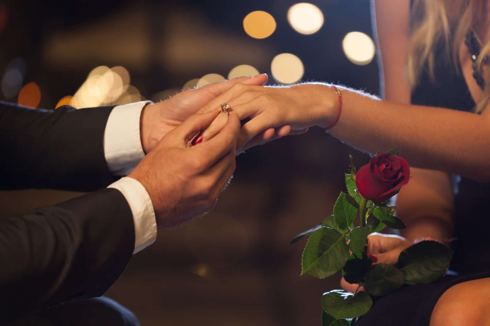 A person kneels to propose, placing a ring on the hand of their partner, who holds a red rose. A person kneels to propose, placing a ring on the hand of their partner, who holds a red rose.
