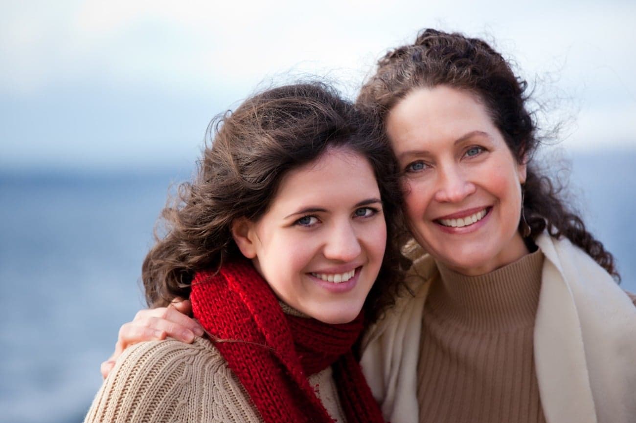 A young woman and her mother smile warmly together against a serene outdoor background. A young woman and her mother smile warmly together against a serene outdoor background.