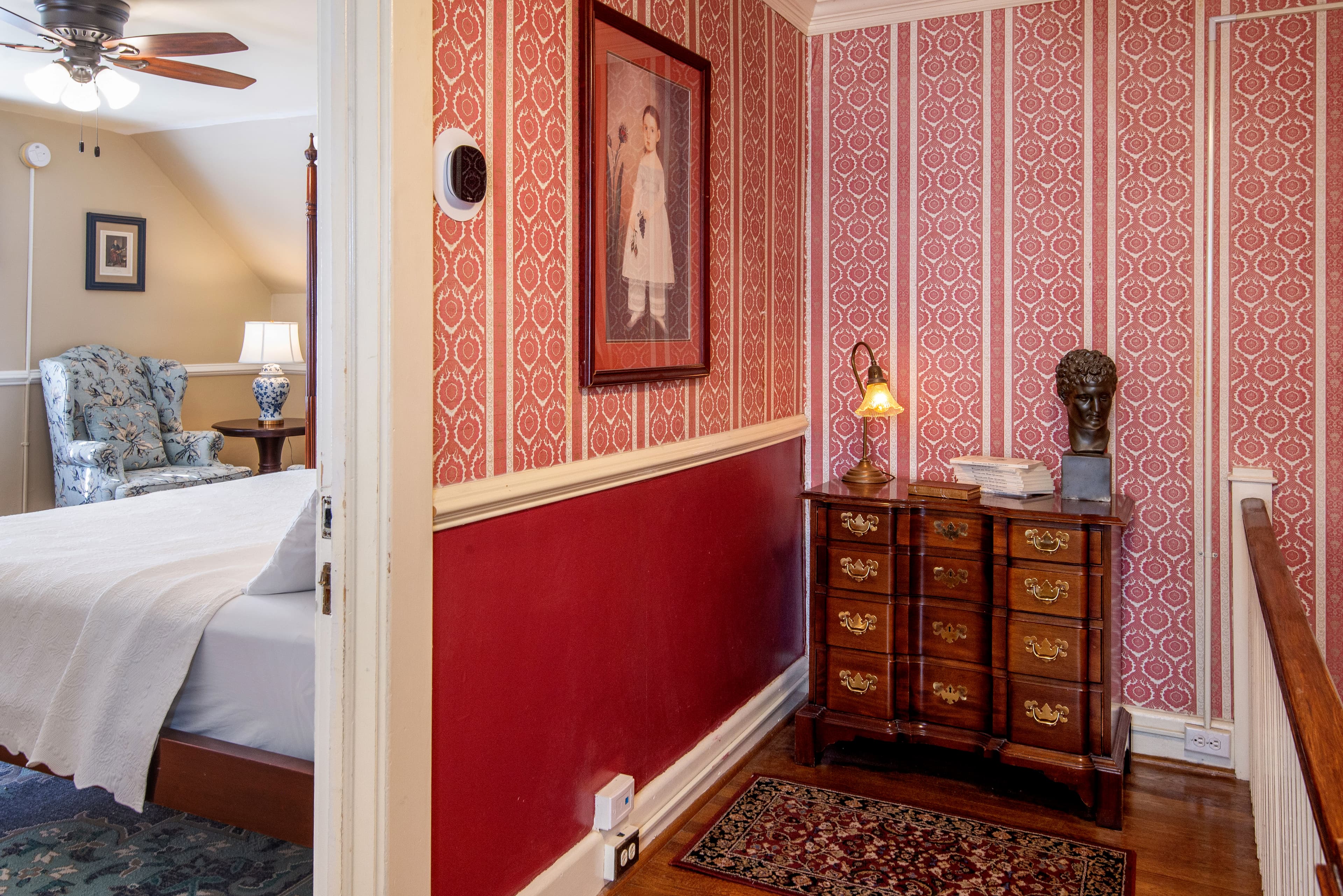 A warmly decorated hallway features red wallpaper, a wooden dresser, and an accommodating view into a cozy bedroom.