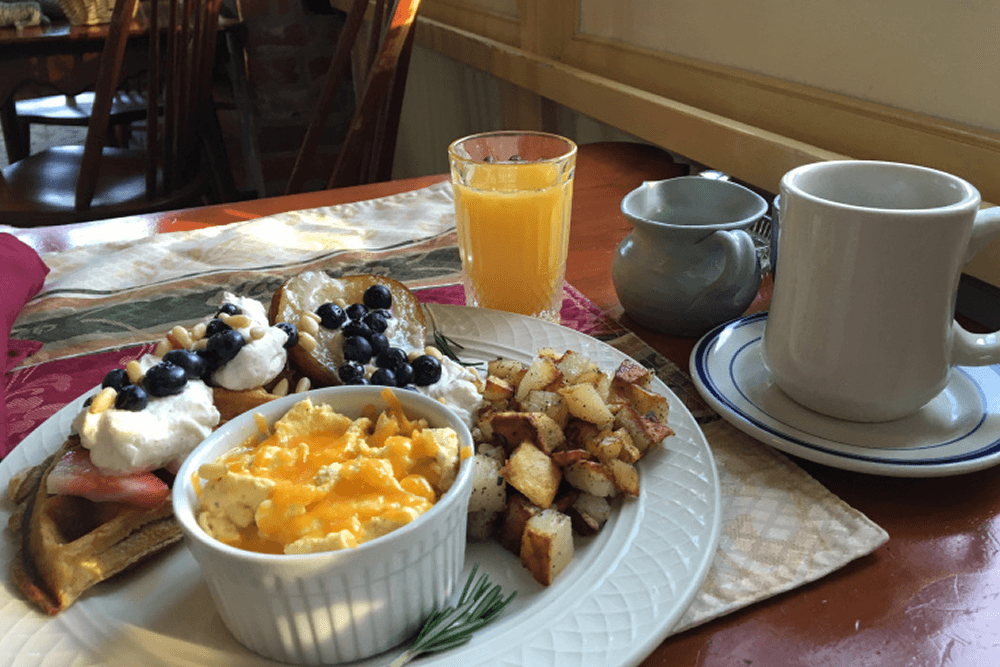 A delicious breakfast spread featuring fluffy waffles topped with berries and whipped cream, a ramekin of cheesy scrambled eggs, crispy hash browns, a glass of orange juice, and a steaming cup of coffee.