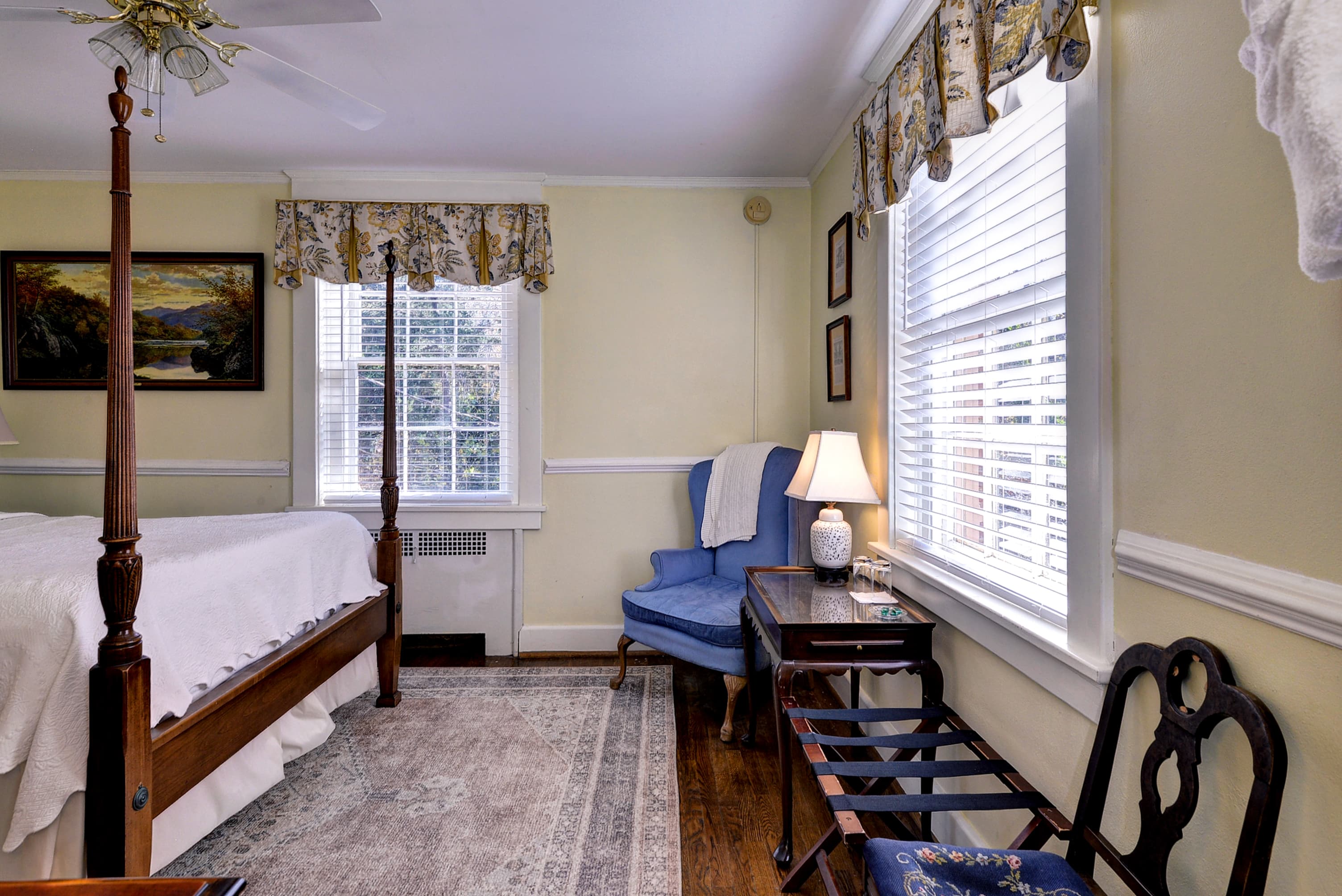 A cozy bedroom featuring a four-poster bed, a blue armchair, and natural light from windows adorned with floral curtains.