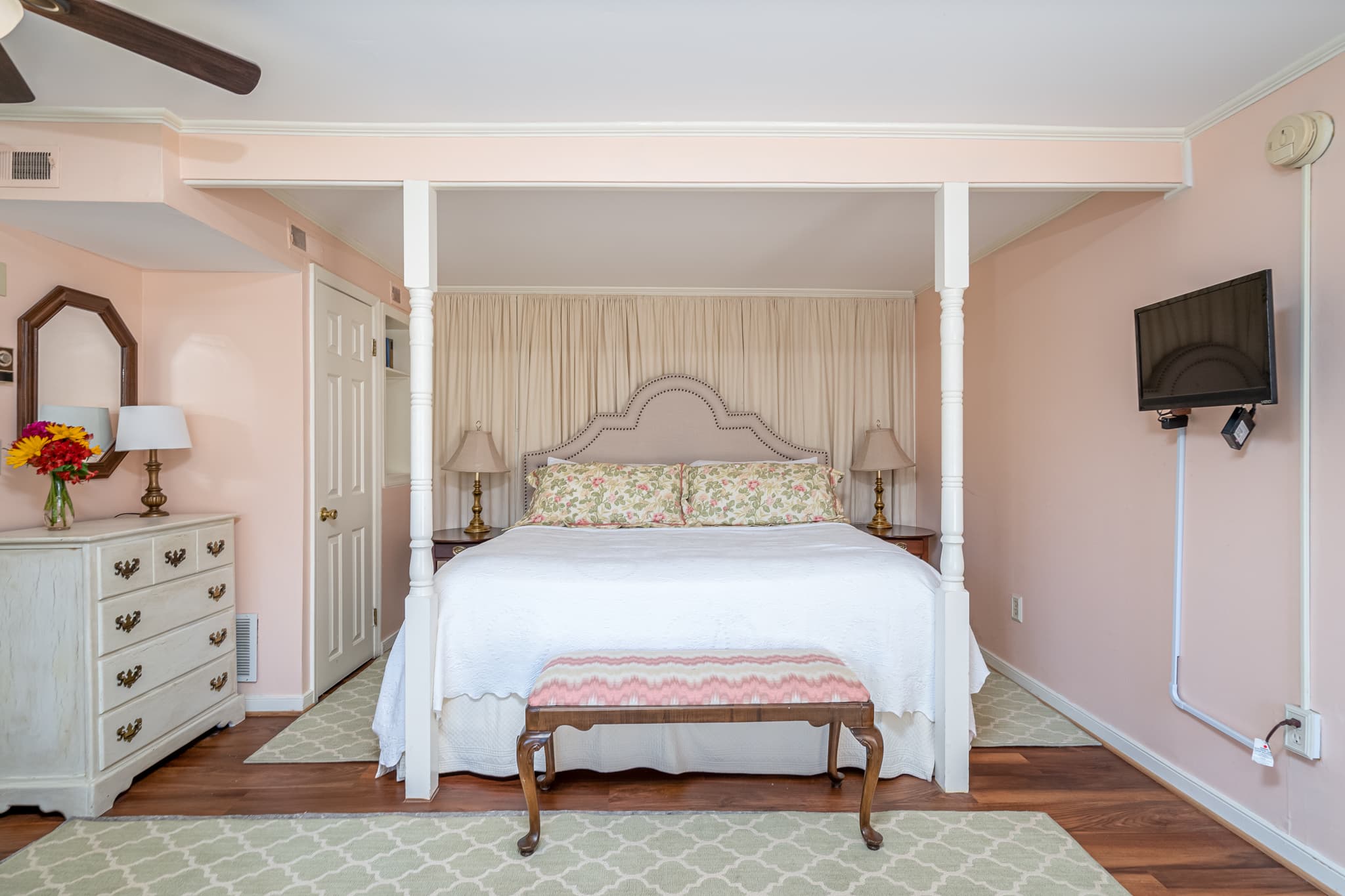A cozy, pastel-colored bedroom featuring a queen-sized bed, a decorative bench, a wooden dresser, and a wall-mounted TV.