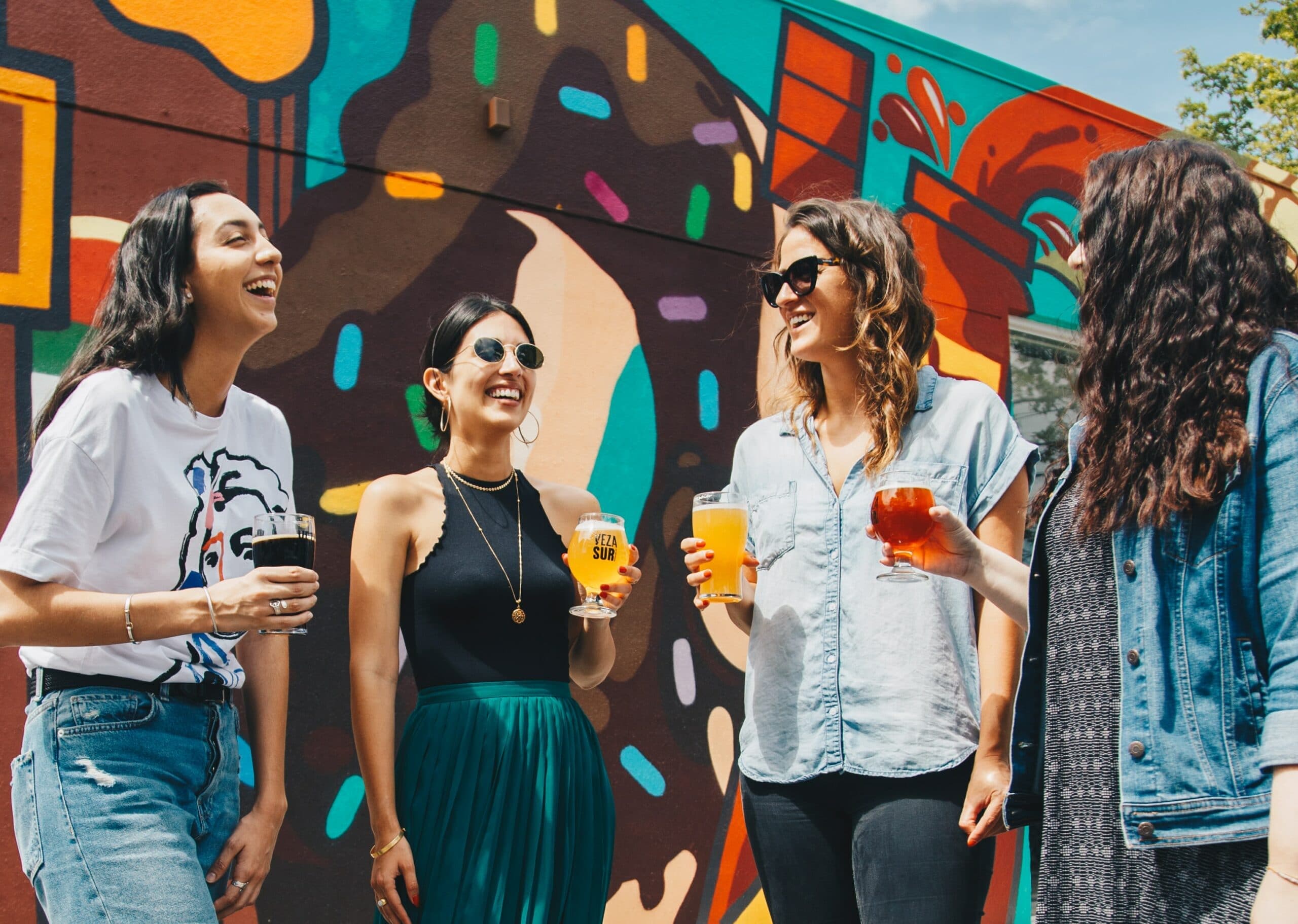 Four women are laughing and enjoying drinks in front of a colorful mural. Four women are laughing and enjoying drinks in front of a colorful mural.
