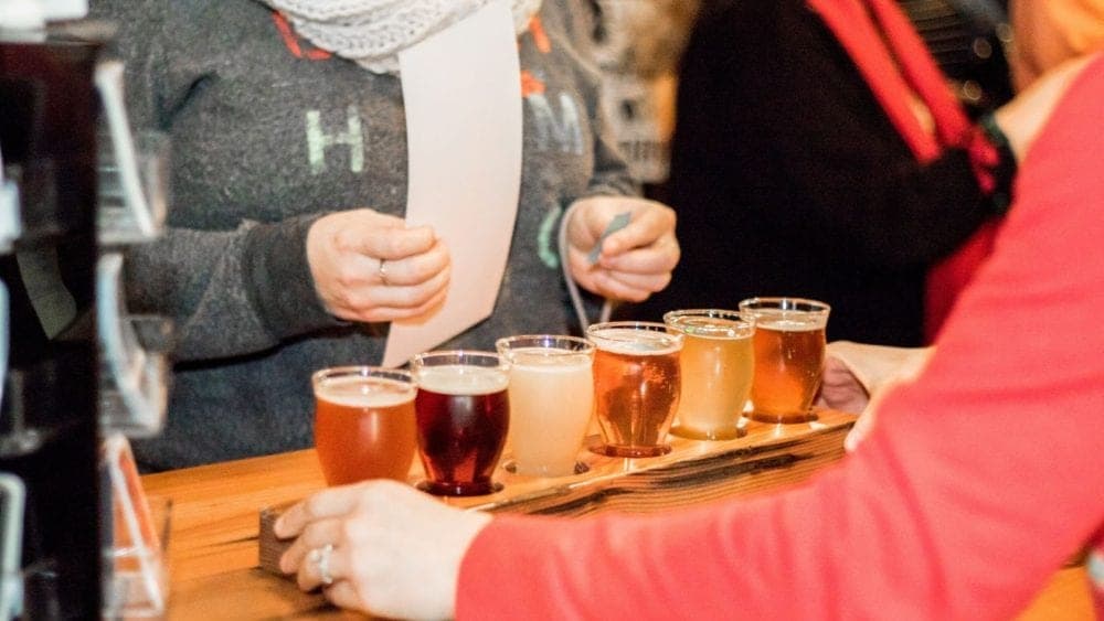 A person is holding a flight of six different craft beers on a wooden paddle at a bar. A person is holding a flight of six different craft beers on a wooden paddle at a bar.