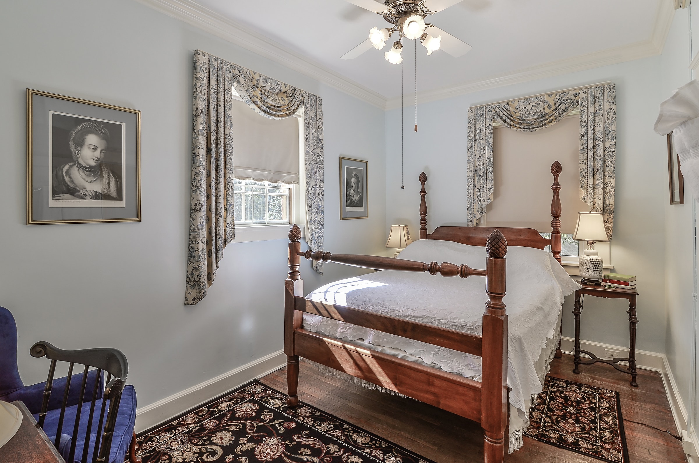Bedroom with four-poster queen bed, richly colored oriental area rugs, nightstand with lamp, ceiling fan