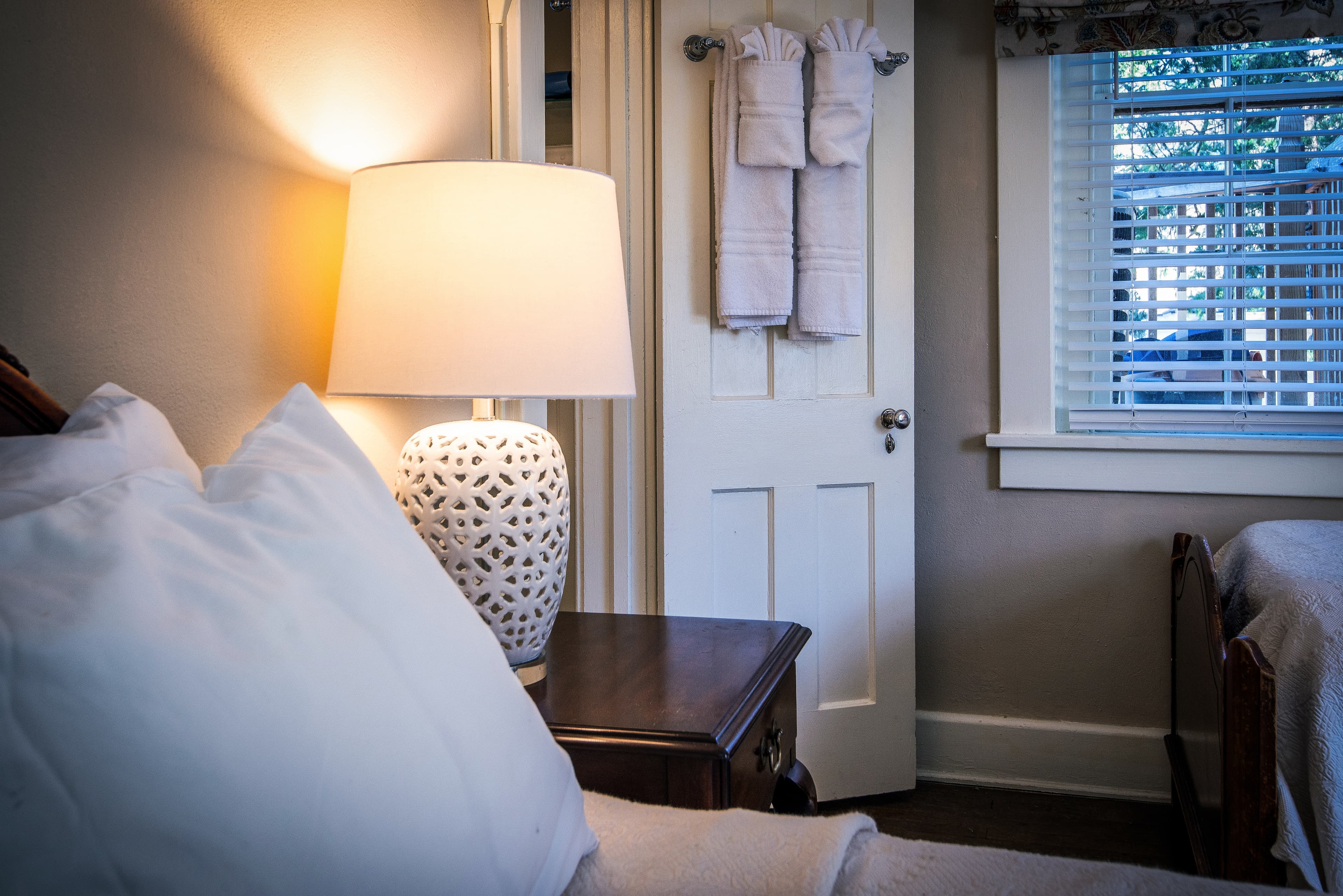 Cozy bedroom interior featuring a bedside lamp, a wooden nightstand, and a door with towels hanging nearby.