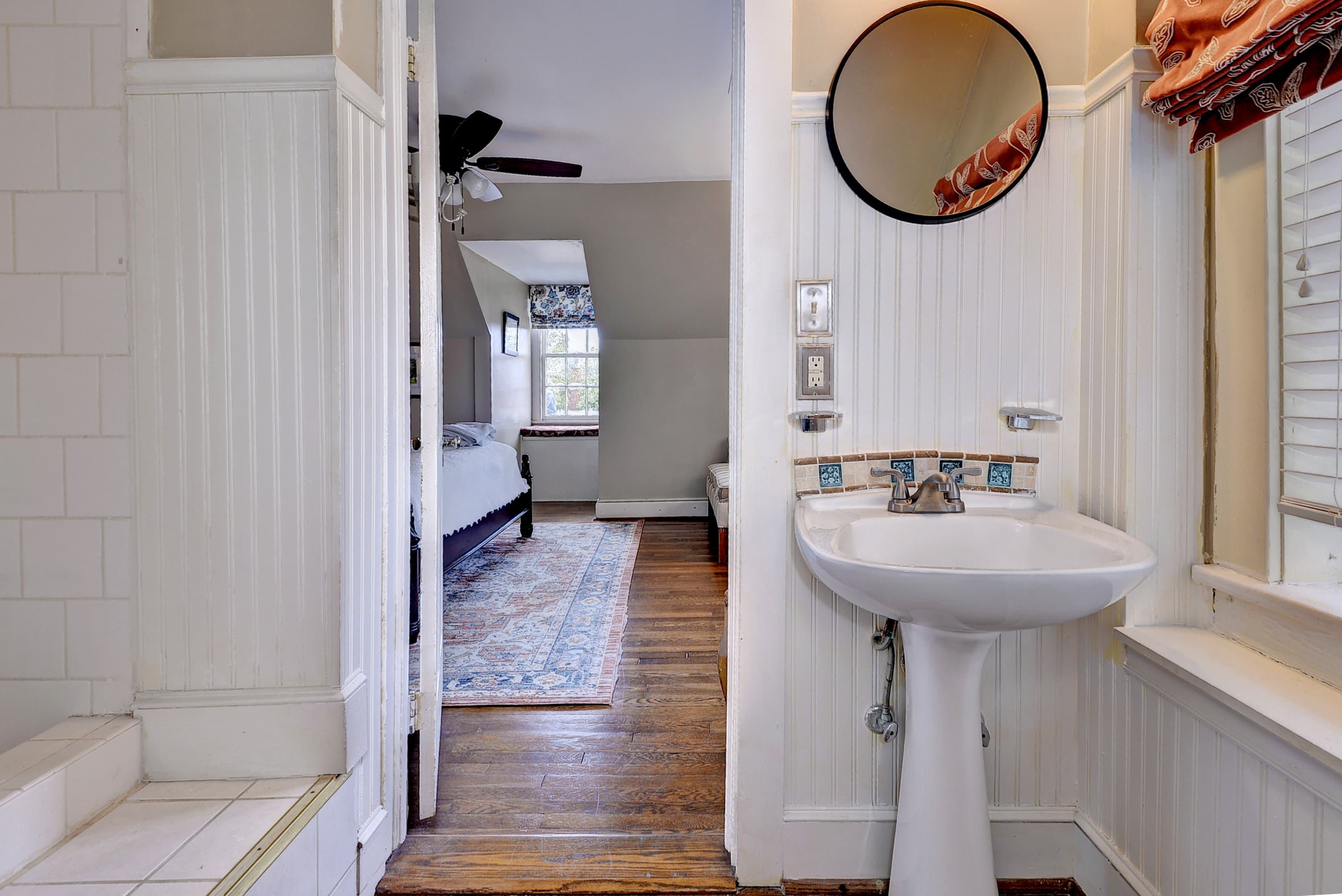 A bright bathroom with a pedestal sink and circular mirror, leading to a cozy bedroom visible in the background.