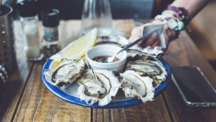 A hand reaches for a plate of oysters with lemon and dipping sauce on a wooden table. A hand reaches for a plate of oysters with lemon and dipping sauce on a wooden table.