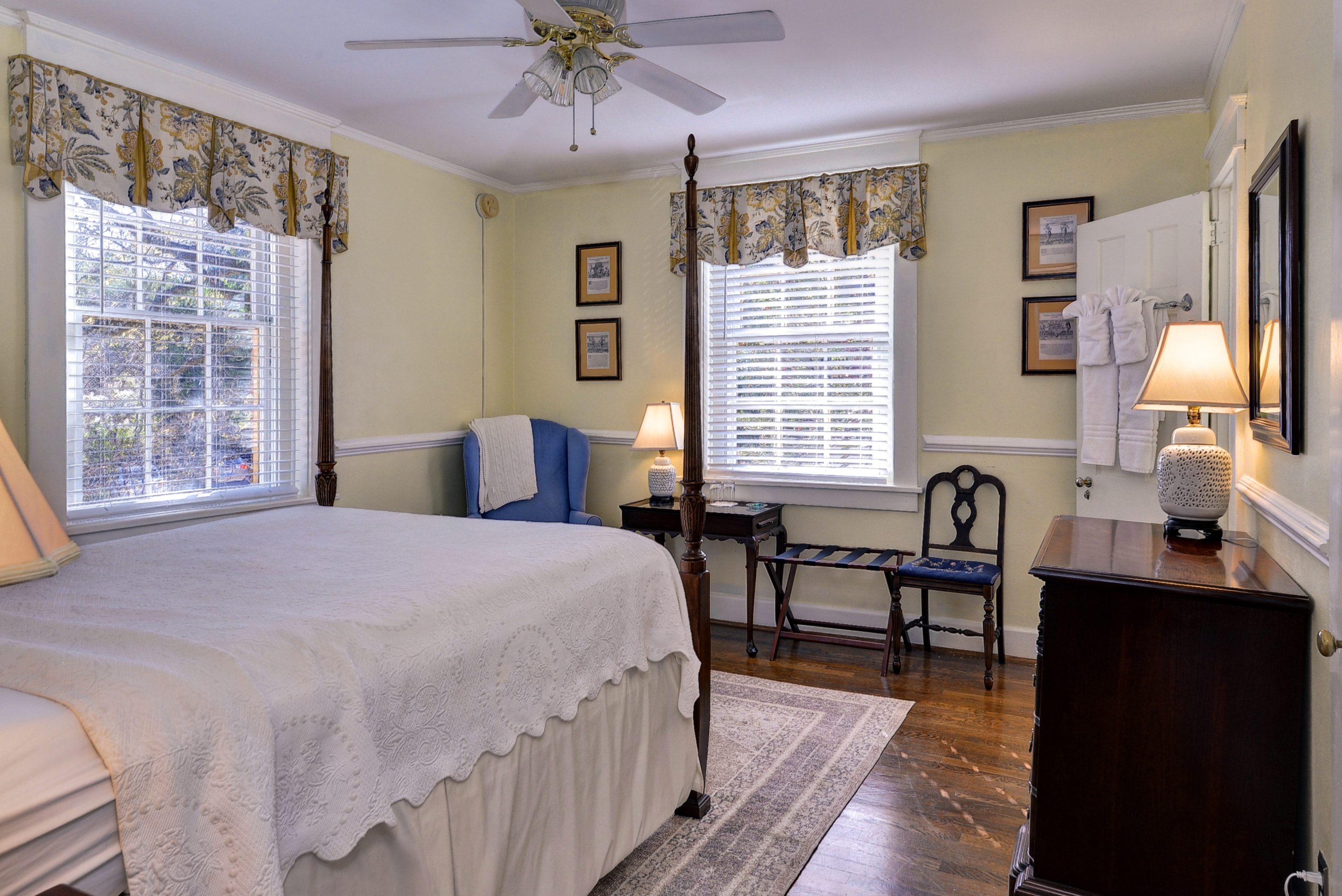 A cozy bedroom featuring a bed, two windows, a chair, and a small desk with warm lighting.