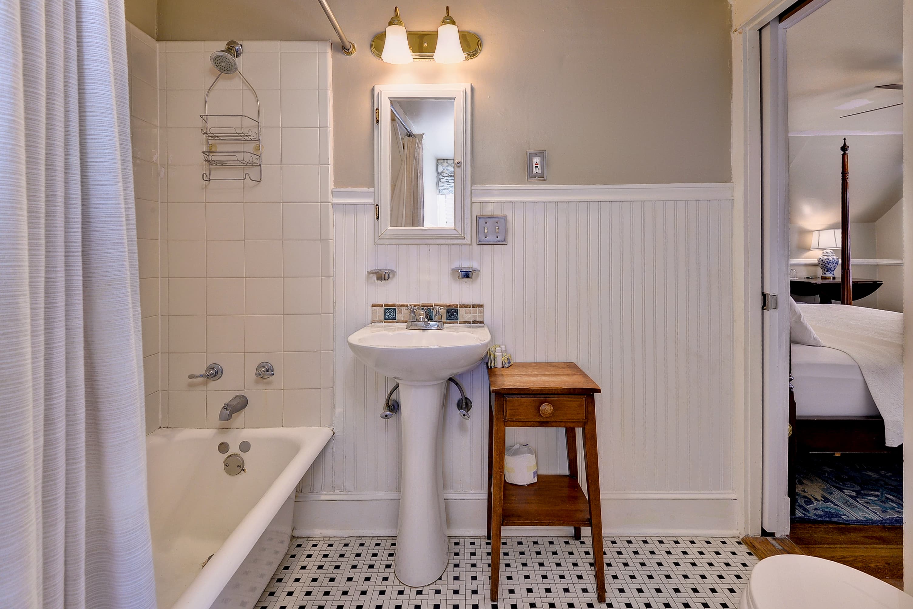 A small, vintage bathroom featuring a bathtub, sink, mirror, and a wooden side table.