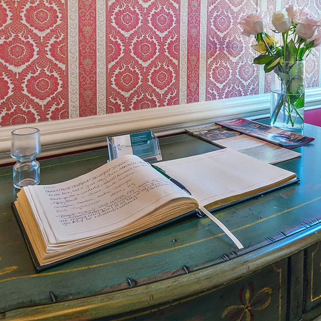 Interior photo of a table adorned with guest information, a hard bound guest book, pen, and vase filled with freshly cut roses.