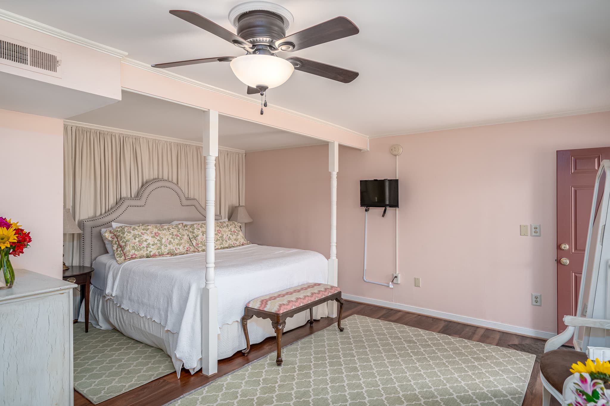 Bright bedroom featuring a king-sized bed, decorative pillows, and a ceiling fan, with light-colored walls and wooden flooring.