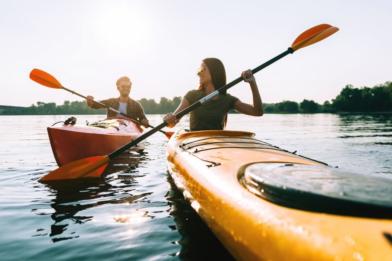 A man and woman paddle kayaks on a calm lake under a bright sky.