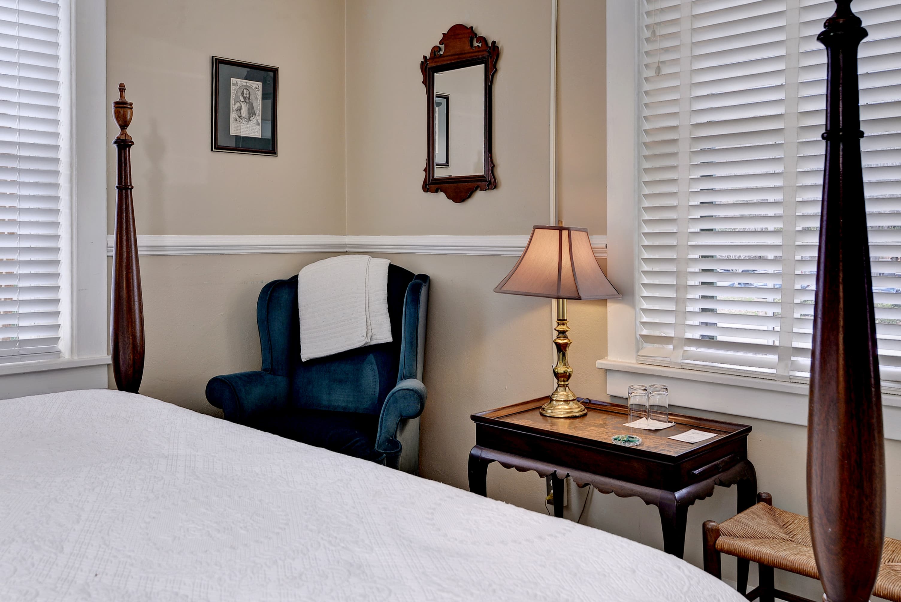 A cozy bedroom featuring a bed, a blue armchair with a blanket, a bedside lamp, and a small table with glasses.
