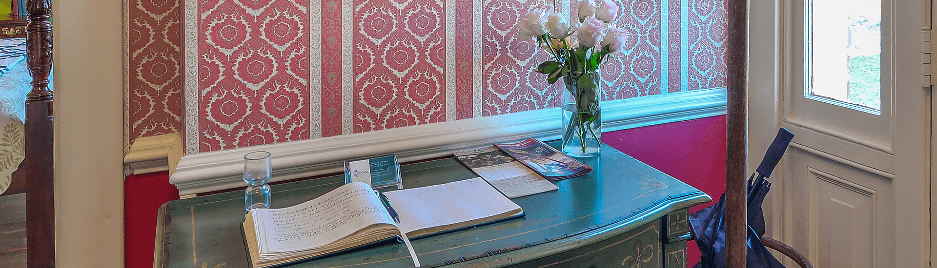Antique front desk table with a vase of roses and a sign in book on top, with floral rose wallpaper on wall behind it.