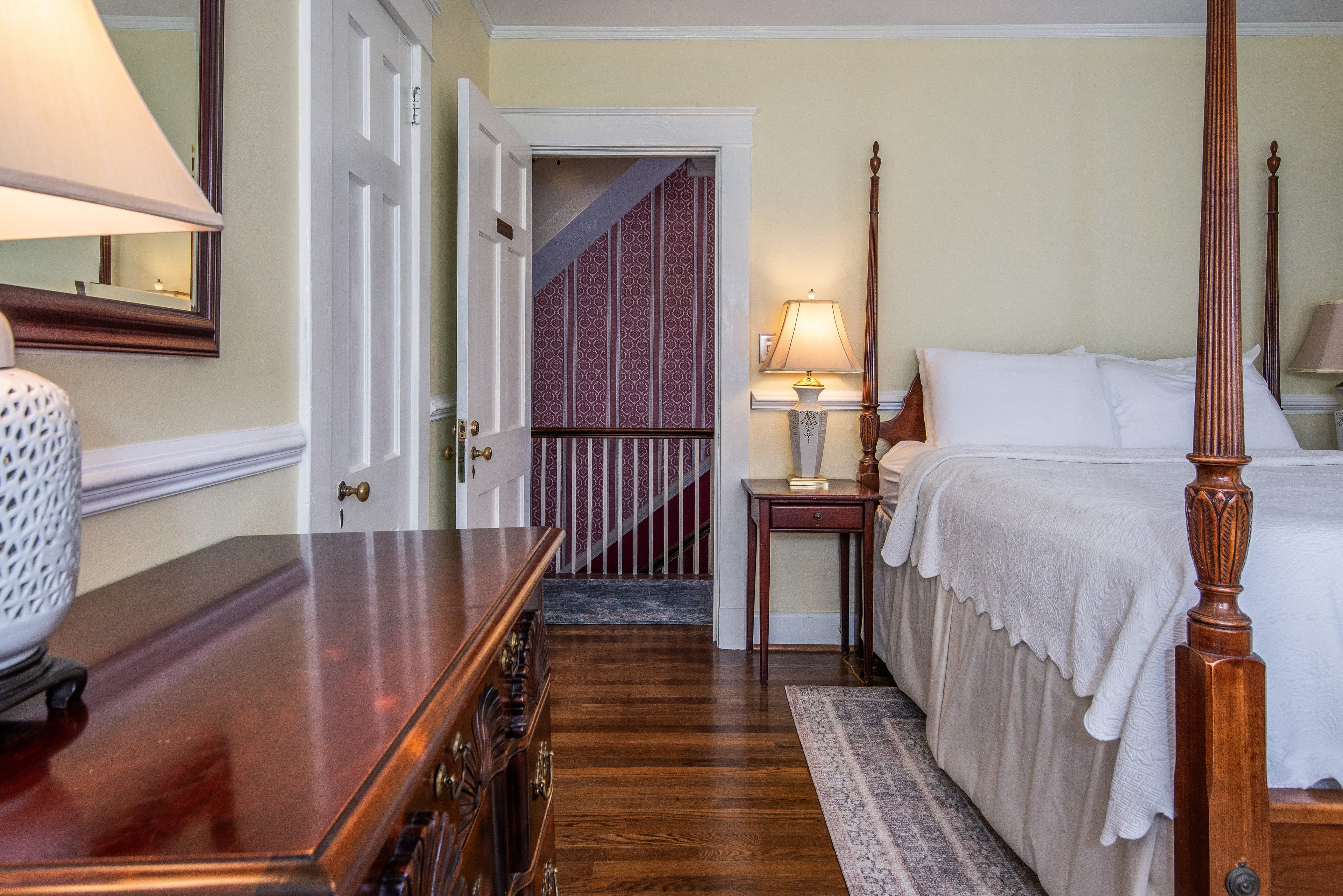A cozy bedroom featuring a four-poster bed, dresser, and a staircase in the background.