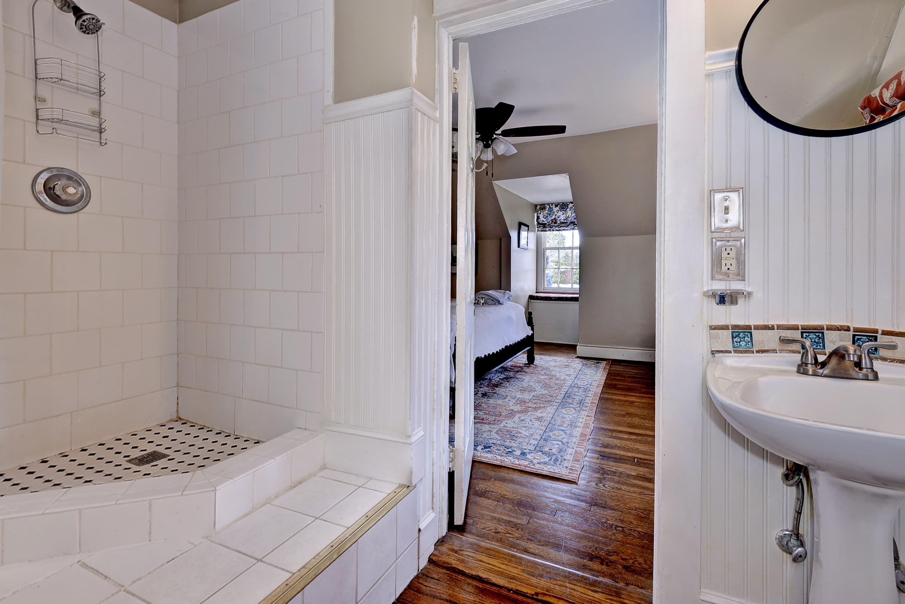 A bright bathroom featuring a tiled shower and a view into a cozy bedroom with hardwood floors.