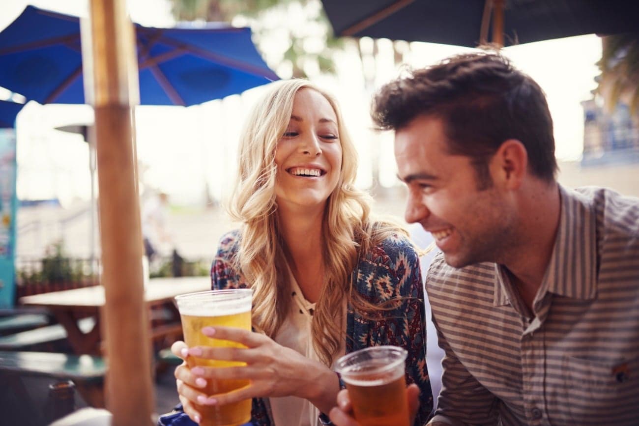A woman and a man sit together at a table, smiling and holding drinks in a relaxed outdoor setting.