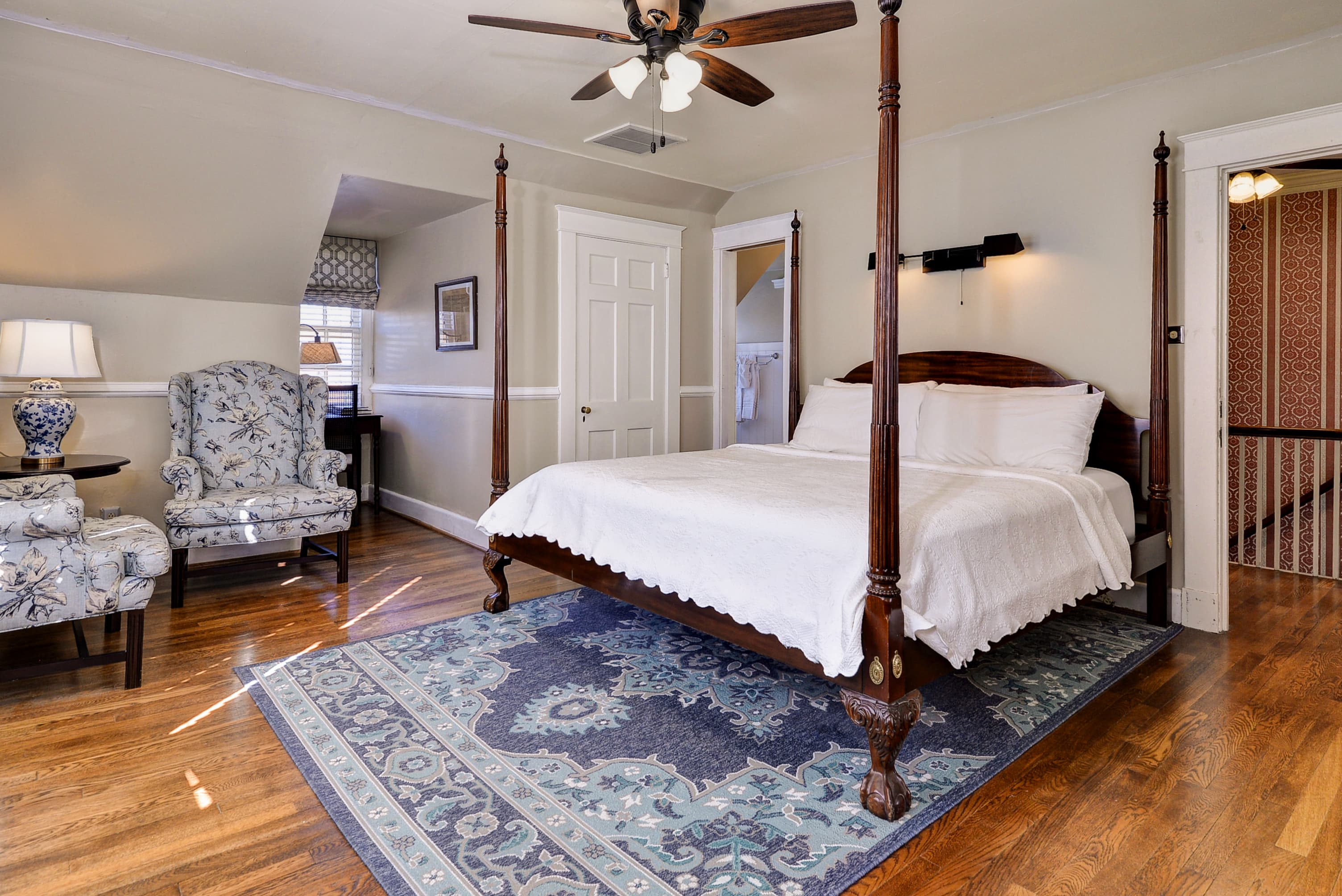A cozy bedroom featuring a four-poster bed, floral armchairs, and a patterned rug on hardwood flooring.