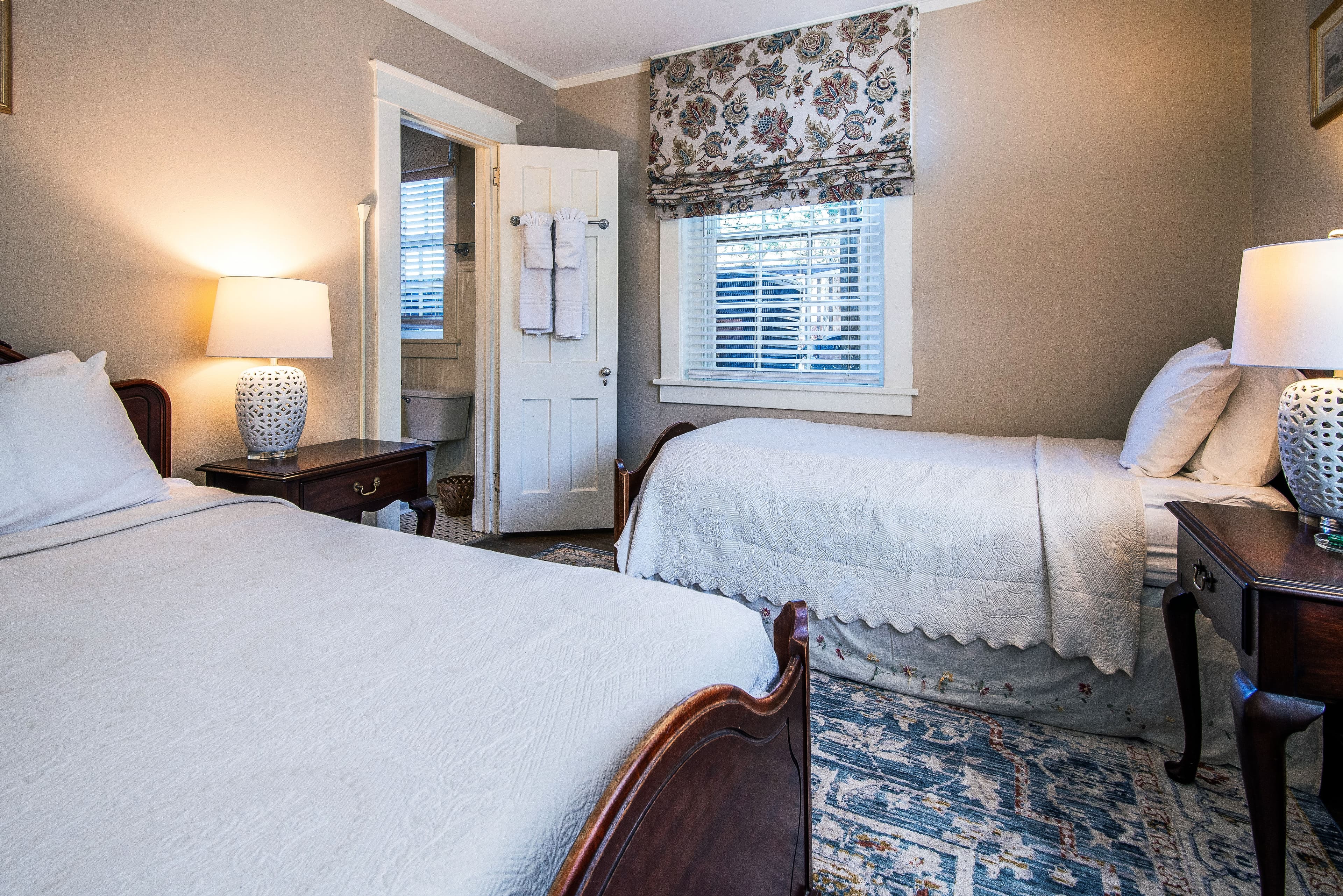 A cozy bedroom featuring two beds, a bedside table with a lamp, and a window with floral drapes.