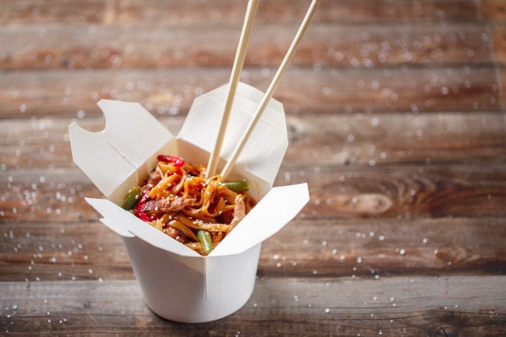 A takeout box filled with noodles and vegetables, accompanied by chopsticks. A takeout box filled with noodles and vegetables, accompanied by chopsticks.